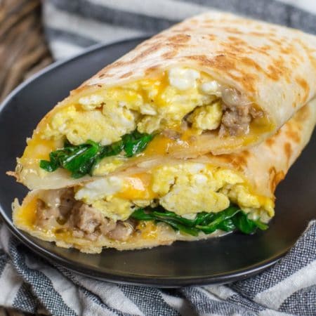 A healthy breakfast burrito stuffed with turkey sausage, spinach, and cheddar cheese cut in half on a black plate.
