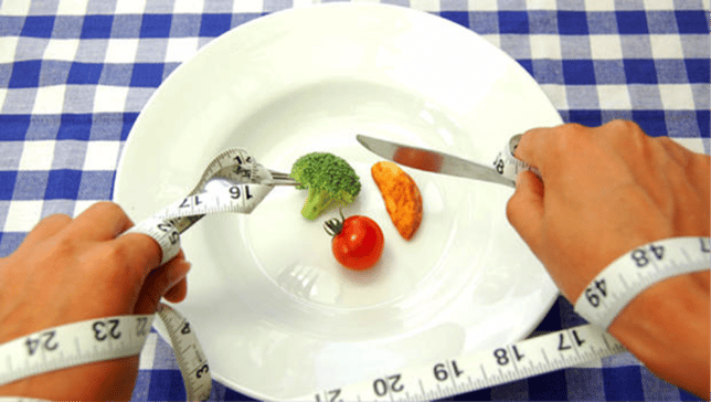A white plate with a single piece of broccoli, a wedge of potato, and a grape tomato with hands holding silverware and wrapped in a measuring tape hovering over it representing the common assumption that diets are restrictive and do not have sustainable nutrition.